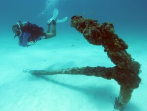 A scuba diver swimming over sand next to a historic anchor.